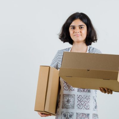 Little girl holding cardboard boxes in t-shirt, apron and looking cheery. front view.